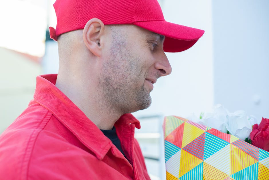 A male delivery person wearing a red cap and matching red jacket is holding a colorful gift bag decorated with geometric stripes in yellow, blue, red, and white, containing a bouquet of flowers. The b