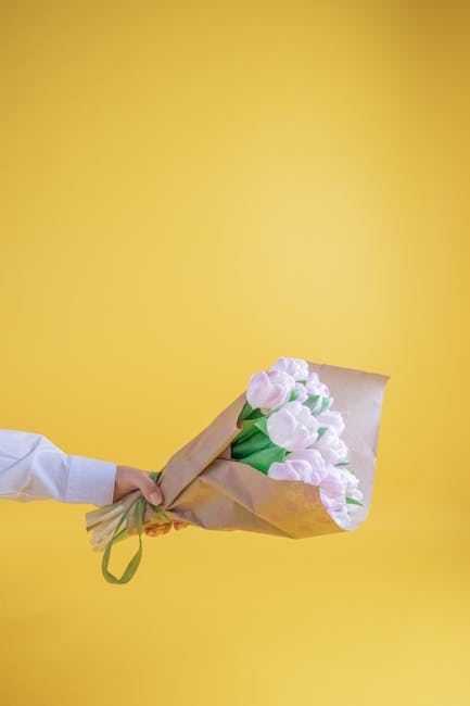 A person wearing a white shirt is holding a bouquet of fresh, pale pink tulips wrapped in brown craft paper with a green ribbon, against a bright yellow background. The tulips have smooth, rounded pet