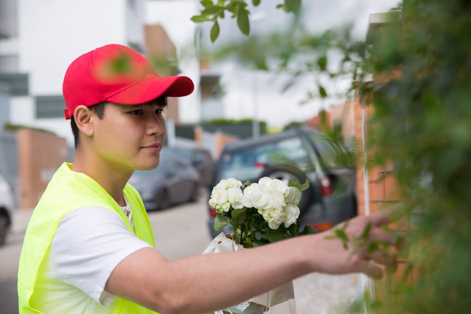 A young delivery person wearing a red cap and high-visibility yellow vest is placing a bouquet of fresh white flowers, including roses and carnations, into a park of greenery outside a residential bui