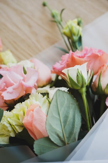 A close-up of a floral bouquet featuring pale pink and cream lisianthus flowers and green leafy foliage, arranged in a white wrapping paper. The bouquet is resting on a light wooden surface, with fres