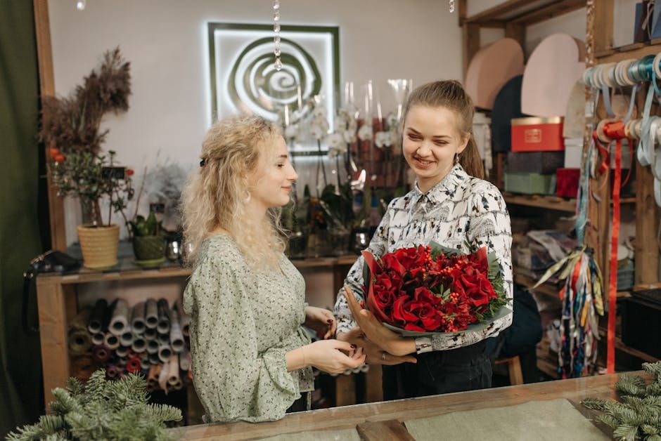 A young woman with long, light brown hair tied back, smiling as she receives a bouquet of fresh red roses, red carnations, and greenery wrapped in brown paper from a florist in a shop. The florist, a 