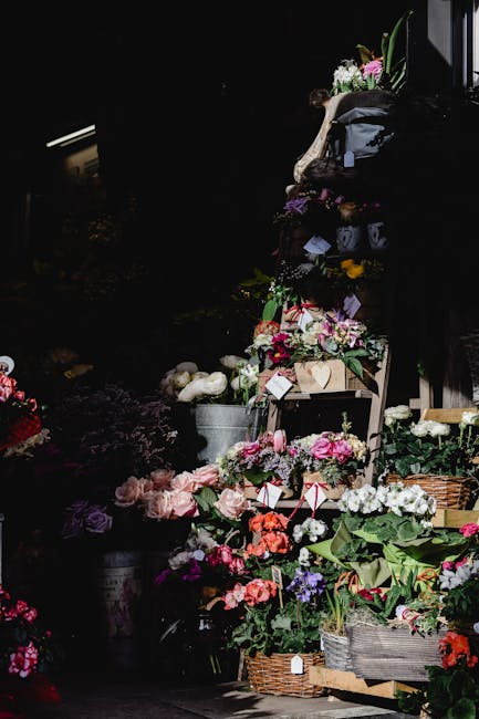 A display of various fresh flower arrangements and potted plants at a floral shop, with vibrant pinks, whites, purples, and reds. The bouquets are arranged in wicker baskets, metal buckets, and wooden
