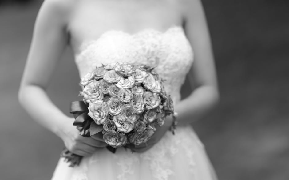 A bride dressed in a strapless lace wedding gown holds a tightly arranged bouquet of small, pale roses with slightly ruffled petals. The bouquet is wrapped with a dark satin ribbon at the base. The br