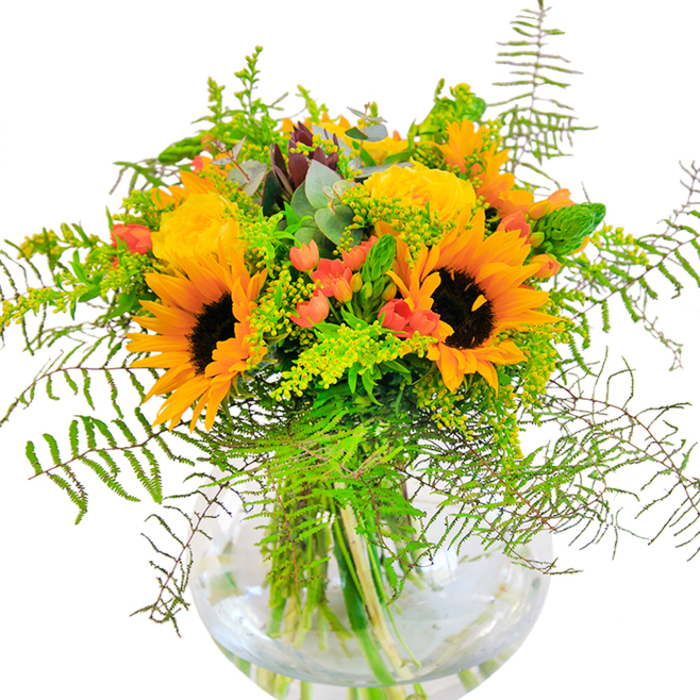 Evoking the uplift of a brilliant morning in Elmers End, this vibrant floral arrangement balances boldness and refinement within a luminous glass vase. The sunflowers' radiant yellow-orange petals and inky brown centres are bright as laughter, forming a cheerful canopy above a spiral of golden yellow garden roses, their tightly clustering petals unfolding with classic elegance. Interspersed among these major blooms, gleaming red hypericum berries provide lively punctuation, their vivid colour a joyful surprise mid-greenery. Airy, featherlike ferns tumble from the bouquet's core, lending gentle movement and drawing the eye downward, while light green fillers tipped with tiny yellow blossoms and slender flower spikes add layers of interest. Soft, bluish eucalyptus leaves bring points of muted coolness and hint at woodland walks through Elmers End Recreation Ground. The transparent round glass vase displays the sturdy green stems, bathed in glistening water, and underscores the freshness and clarity the florist delivers each day. Gently perfumed, the bouquet calls to mind sunlit afternoons and the warmth of community ties. This ensemble perfectly suits celebratory occasions-birthdays, anniversaries, or impromptu gatherings-where a burst of local colour and floral artistry is treasured. Every detail, shaped by expert Elmers End Florist hands, communicates both the beauty of flowers and a sense of local belonging.