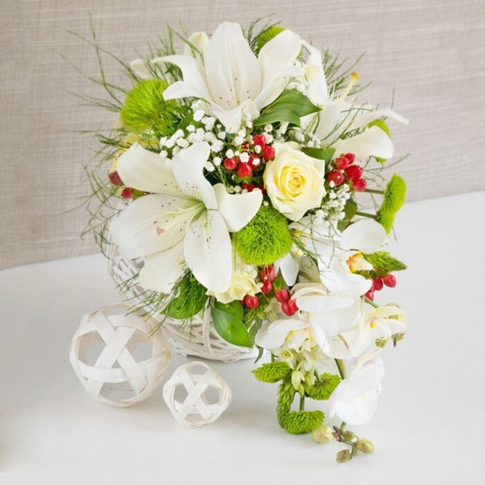 A finely crafted wedding-style centerpiece is arranged on a glossy white tabletop, its cascading structure and soft colour palette evoking elegant celebrations in and around Elmers End. The upper section of the arrangement is dominated by large white Oriental lilies, their trumpet-shaped petals opening wide to reveal speckled interiors and prominent yellow stamens that seem to glow in the gentle light. Close to the centre of these lilies, a single pale cream rose unfolds, its velvety petals cupped tightly together, lending a warm, romantic focus amidst the surrounding white. From the right-hand edge, a stream of white phalaenopsis orchids flows downward in a graceful arc, each orchid bloom composed of smooth, rounded petals with a subtle central accent, creating a waterfall or teardrop effect that adds length and fluidity to the piece. Enveloping the main flowers, an abundance of rich greenery spills outward: fine, feathery fronds, slim grass-like blades, and bright green pom-pom flowers that offer a soft, tufted texture. Delicate sprigs of baby's breath are interwoven throughout, their minuscule white blooms adding an airy, cloud-like quality, while clusters of deep red berries are dotted strategically, giving sharp, festive highlights reminiscent of winter walks through nearby Beckenham or along Elmers End Road. All of the stems are anchored securely within a low, round white wicker basket whose woven texture complements the natural theme and keeps the arrangement perfectly suited for a centre of the table. To the left, resting on the same white surface, two decorative white wicker balls of differing sizes echo the basket's material, adding sculptural interest and visual balance. A softly textured beige background frames the entire scene, its neutral tone allowing every detail-the lilies, orchids, rose, foliage, and berries-to stand out clearly, creating an overall impression of purity, grace, and carefully considered floral design.