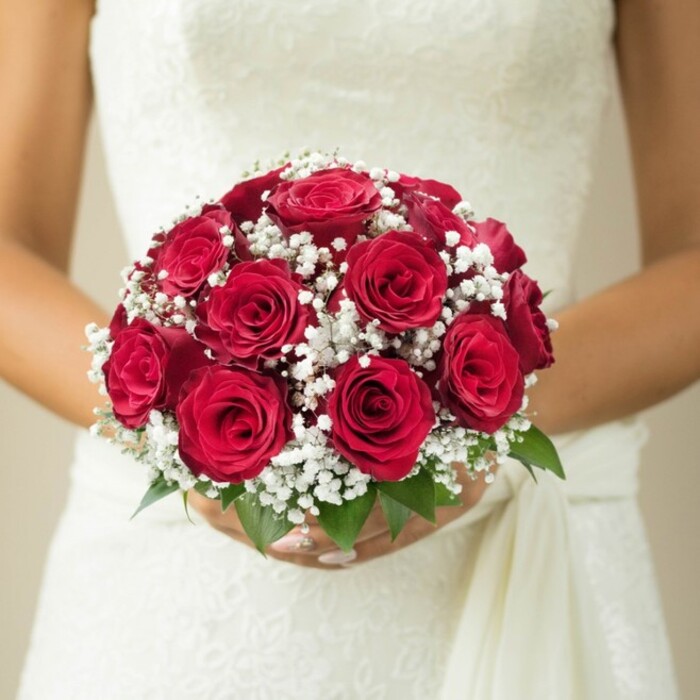 An intimate, mid-torso portrait captures a bride in Elmers End holding a bouquet shaped with loving precision. The arrangement is composed almost entirely of deep red roses, each at its peak, their velvet petals coiled in plush concentric circles that yield a sensation of depth and richness. Among the roses, scattered tufts of white gypsophila provide a gentle, cloud-like softness-a hallmark of timeless wedding florals. Wisps of fresh green leaves peek through at the base, drawing a connection to the leafy parks and family gardens found throughout Elmers End's friendly neighbourhoods. The bride's hands, with their immaculate manicure, encircle the bouquet with care, while the bodice of her wedding dress-possibly adorned in lace or subtle embroidery-acts as a backdrop, catching soft daylight. A white draping sash falls to one side, offering fluidity and a sense of anticipation. The softly blurred cream-toned background keeps the visual focus firmly on the artful interplay between flowers and fabric. Crafted with a florist's attention to detail and a neighbourly warmth, this image embodies the elegance and heartfelt tradition that define weddings and special occasions throughout Elmers End.