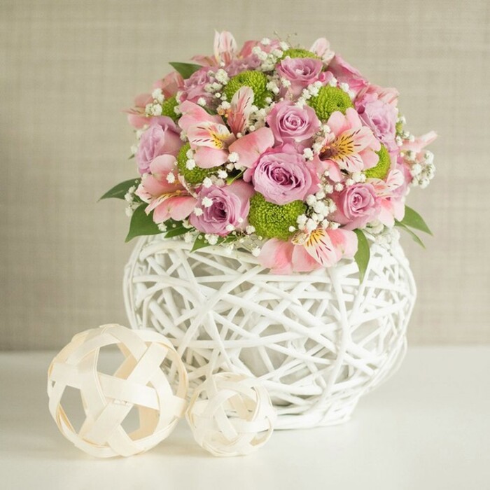 Centered on a spotless white surface in Elmers End, this ornate floral centerpiece radiates serenity and refined artistry. Full, pastel pink and lavender roses form the heart of the arrangement, each blossom revealing a velvety softness. Interspersed are sprigs of elegant pink alstroemeria, whose delicate, streaked petals introduce subtle movement and depth, their trumpet-shaped blooms glowing with bright highlights. Energetic pops of lime green button chrysanthemums nest within the bouquet, their compact, spherical forms providing playful intrigue-reflecting the lively natural beauty found along Elmers End's leafy streets near the local market. Dainty white baby's breath fills in the gaps, lending an ethereal, floating effect, while deep forest green leaves peek between the blooms for grounding contrast. The ensemble rises from a distinctive, lattice-style vase: crafted from open, intricately interwoven white strips, the vessel's rounded shape and modern pattern echo innovative design trends in Elmers End's creative community. On the left side, two decorative balls, woven from creamy, rattan-like fibers, complete the visual story-one markedly larger, the other a bit forward, both accentuating the vase's openwork motif. The background, a gentle beige with fabric-like texture, frames the scene gracefully, inviting the colors and textures to pop. Perfect as a wedding table centerpiece, housewarming gift, or a gesture of heartfelt celebration, this arrangement-rooted in Elmers End Florist craftsmanship-perfectly marries natural beauty with local, contemporary artistry.