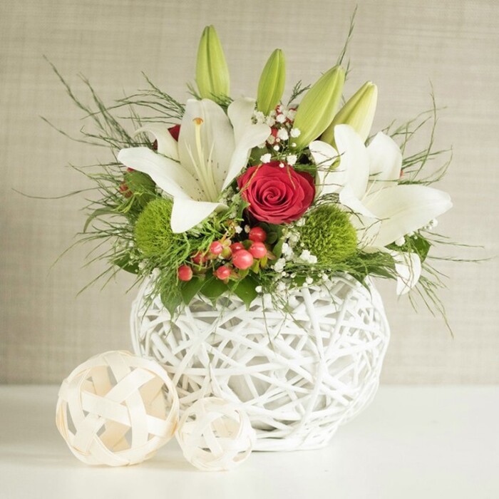 A refined, garden-fresh centerpiece sits prominently on a white surface, imagined as the focal point of a wedding table or intimate celebration in Elmers End. The arrangement is designed in a round white vase with a distinctive open-weave texture, as if slender twigs or white rattan have been gently woven into a perfect orb. From this airy base, white lilies reach upward and outward, some fully open with broad, silky petals curling back to reveal speckles and warm yellow stamens, others still enclosed in smooth green buds that lend vertical interest and convey freshness. At the heart of the composition is a single red rose, its tightly layered, velvety petals forming a rich crimson rosette that immediately catches the eye and introduces a romantic, almost classic bridal tone. Around it, bright green pompon-style blooms cluster like soft cushions, resembling green dianthus or chrysanthemums and bringing a vivid, contemporary twist to the traditional red-and-white theme often seen in Elmers End wedding venues. Fine sprays of white baby's breath are threaded between the main flowers, adding delicate, cloud-like detail that softens the transitions between blooms. Small clusters of glossy red berries peek out from the greenery, offering a subtle nod to festive seasons without overpowering the design. The whole arrangement is framed by a generous mix of foliage: slender, feathery fronds arc outwards with a sense of movement, while deeper green, broader leaves fill in the gaps and give a lush, full silhouette. To the left, two decorative spheres made from pale woven strands echo the vase's pattern and balance the composition. Against a softly textured neutral wall, the arrangement's colours and textures stand out beautifully, capturing the elegance of a locally crafted Elmers End table arrangement.