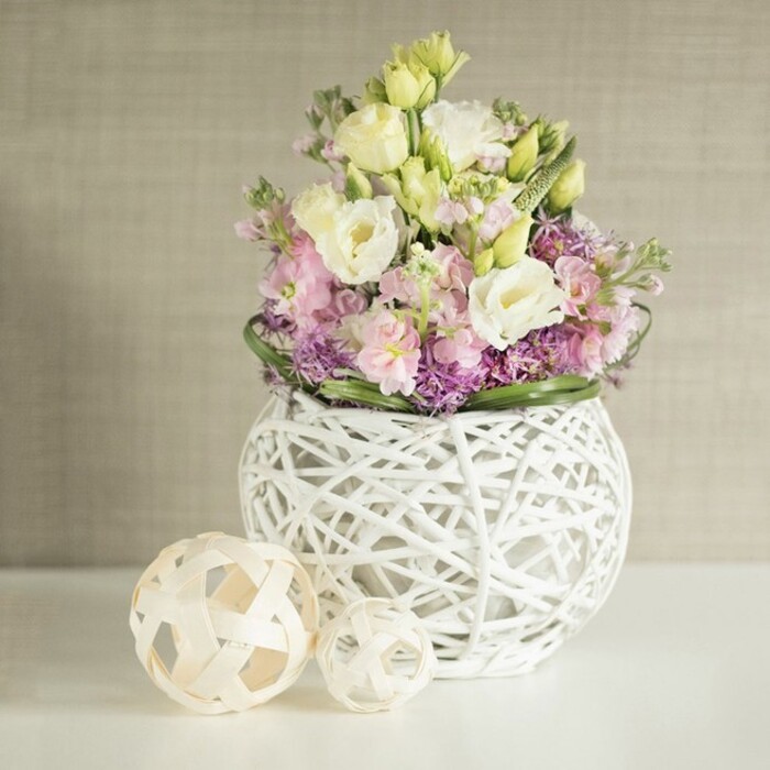 Captured in delicate detail, this floral arrangement is a testament to the botanical artistry found at Elmers End Florist, just steps from the heart of Beckenham. A spherical white vase-formed by a lattice of slender wooden or rattan strips-anchors a bouquet abundant with creamy white and soft yellow lisianthus, their layered, ruffled petals providing delicate movement. Rising through their midst, vivid pink stock flowers lend a vertical grace that guides the eye upward, while nestled below, tiny violet filler blooms (possibly asters or petite alliums) generate dimension and rich texture. The arrangement is encircled by sleek, deep green foliage, echoing the lushness of Elmers End's tree-lined streets and garden spaces. To the side, two woven orbs-crafted from pale, natural materials-offer artisanal harmony to the centerpiece and evoke the creative spirit of local flower stalls near Elmers End's railway station. The composition is set against a subtly textured beige background, reminiscent of sunlit living rooms across the area, where soft diffused light brings out the gentle contrasts between flower and vessel. Designed for lasting impact, this bouquet conjures feelings of freshness, tranquility, and connection to nature, making it a standout choice for a wedding table, spring celebration, or as a radiant focal point for a home styled with inspirations from Elmers End's blend of tradition and modernity.