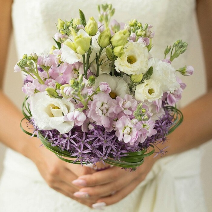 Illuminated by gentle daylight, this captivating bridal bouquet is held with care just above a softly blurred ivory wedding dress, the dappled lace of the fabric visible in the image's lower background. Designed by a local Elmers End Florist, the composition seamlessly blends creamy white Lisianthus in full bloom, each ruffled petal encircling a green-centred heart-symbols of purity artfully paired with slender, unopened buds. Cloud-like clusters of light pink and subtle lilac stock add rich texture and an understated scent evocative of springtime walks along Clock House Road. At the base, a dramatic band of spiky, purple Allium creates a bowl of contemporary edge and depth, their architectural stars grounding the more romantic silhouettes above. Slim, glossy loops of bear grass wrap elegantly around the bouquet's center, contributing lively green structure that echoes the lushness of Elmers End's meadows. The bouquet rests in hands finished with nude or pale rose manicure, while the background's soothing blur ensures attention remains on the vivid, fresh arrangement. Every element speaks to a modern yet timeless approach to wedding floristry-ideal for local brides seeking both tradition and innovation in celebration.