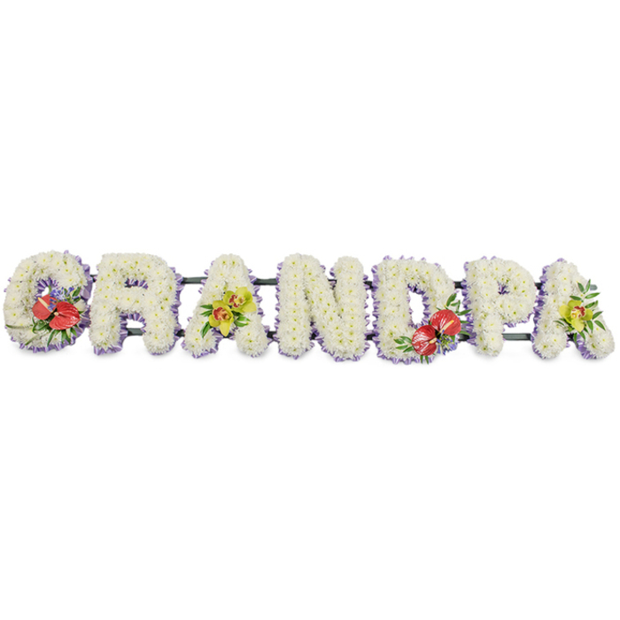 A personalised funeral flower tribute spelling the word GRANDPA stretches horizontally across the image, poised against a bright white background that throws all the colours and textures into clear relief. The letters are bold and blocky, each one built up from a thick covering of small, pristine white chrysanthemums, packed closely together so that no gaps of frame or foam show through. This dense layer of petals gives a soft, quilted appearance, almost like a cushion or pillow crafted entirely from flowers, suggesting rest, comfort, and peace. Along the base of every letter runs a slim trim of pale lilac or lavender ribbon, just visible as a gentle band that traces each outline with cool pastel tones, adding a refined finish without overwhelming the purity of the white blooms. The letters sit securely on a dark green rail that connects them, designed so the tribute can be displayed on a hearse, placed in the chapel, or rested graveside at funerals around the Elmers End area. Four letters feature distinct accent sprays that break the white with meaningful colour: the G and D each display a striking cluster of deep red anthuriums, their glossy, heart-shaped spathes and central yellow spadices shining against rich green foliage and flecks of purple filler flowers. The first and last A are softened with yellow-green orchids, their delicate, exotic blossoms and lush leaves bringing a touch of elegance and the suggestion of ongoing life. This GRANDPA tribute feels thoughtful and deeply affectionate, the kind of bespoke piece created by experienced local florists in Elmers End to honour a dearly missed grandfather.